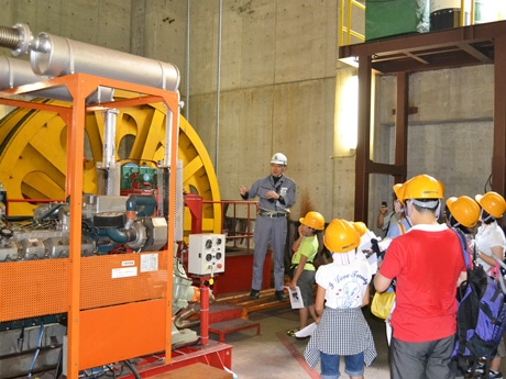 機械室の巨大な原動機を前に整備職員の説明を聞く子供たち