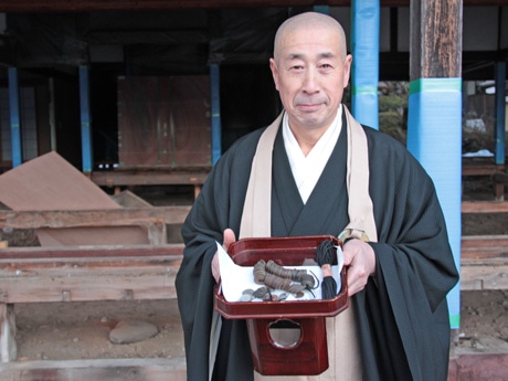 改修中の本堂（写真後ろ）で発見された古銭を手にする住職の三木俊岳さん