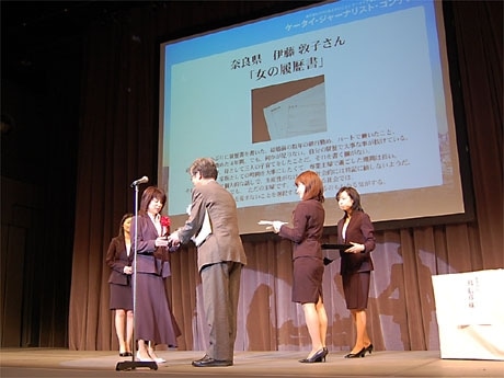 嶌審査委員長からトロフィーを受け取るグランプリ受賞の伊藤さん。スクリーンには伊藤さんの作品が
