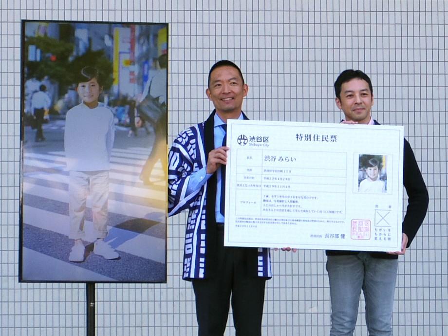 「渋谷みらい」の特別住民票を紹介する長谷部健渋谷区長（左）と井澤友郭こども国連環境会議推進協会事務局長（右）。モニターに映っているのは「渋谷みらい」