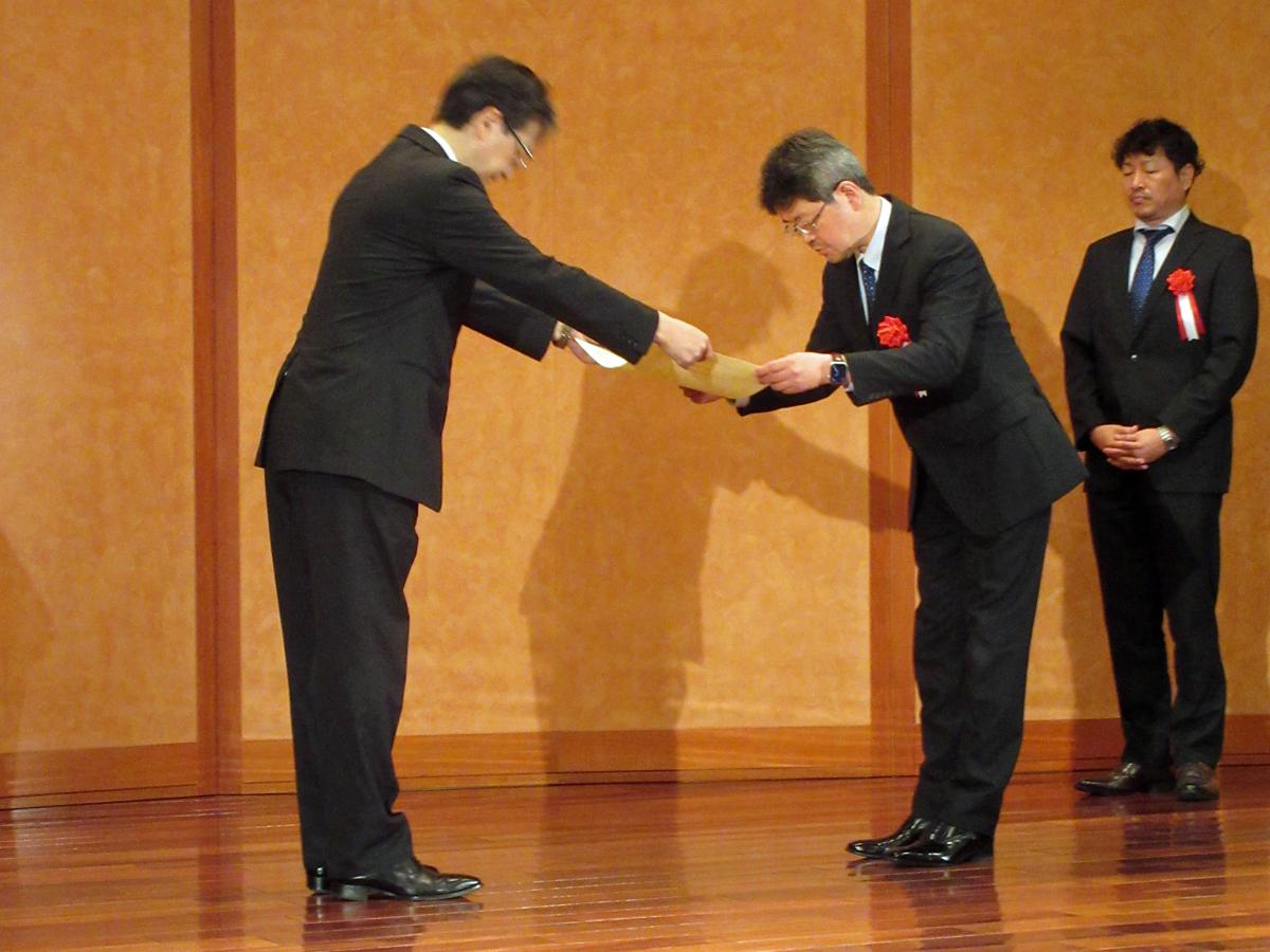 日本都市計画学会の森本章倫会長（左）から賞状を受け取る、渋谷区 まちづくり推進部 渋谷駅中心五街区課の行廣勝哉課長（中央）