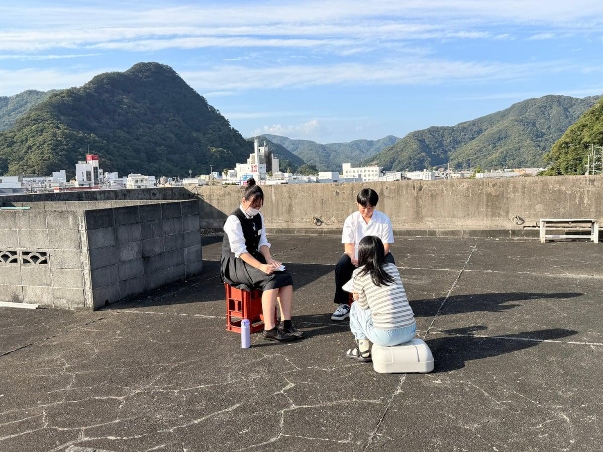 青空と山々を望む屋上で車座になって語り合う高校生たち