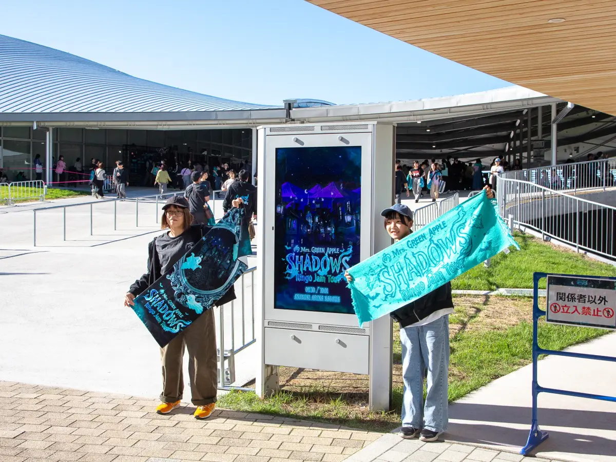 グッズのタオルを広げ、ツアーキービジュアル看板と写真を撮る親子