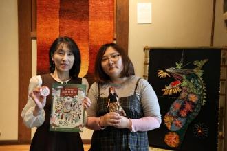 みんなのお家の玄関にある芯アートの横でチラシを手に来場を呼びかける高橋さん（左）と倉田さん（右）